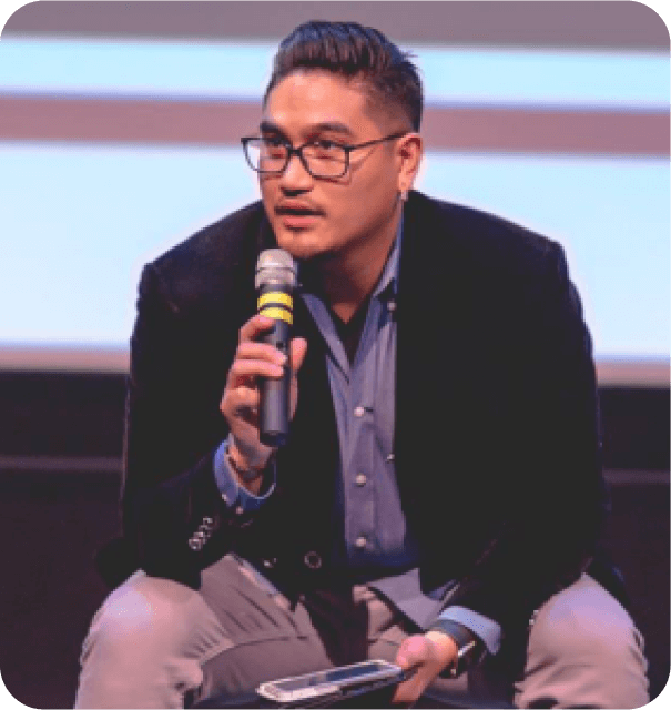 A man wearing glasses is seated on a stage, engaged in a thoughtful expression during a presentation.